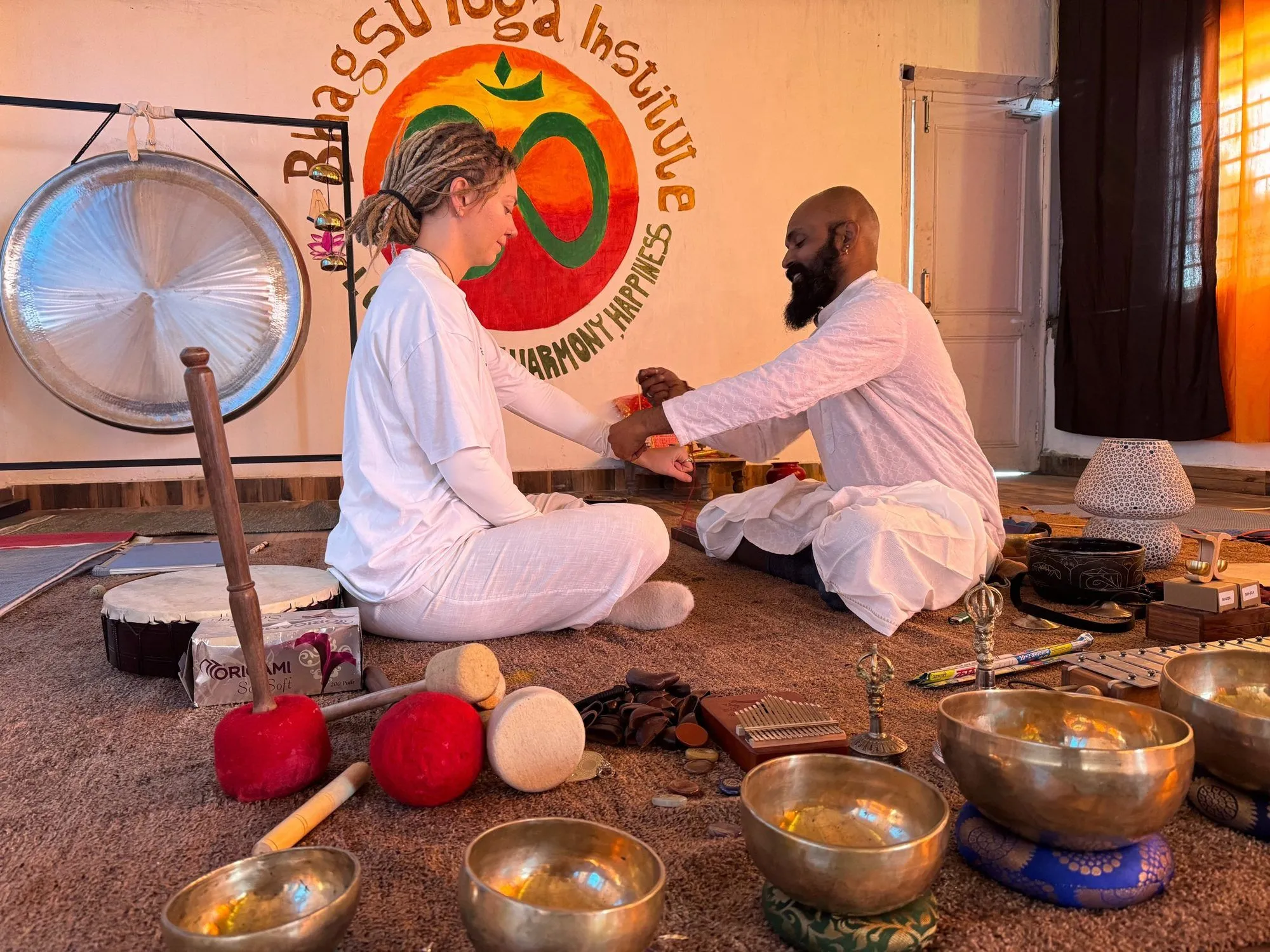 A student and Omji sitting on the floor with a gong and brass singing bowls around them